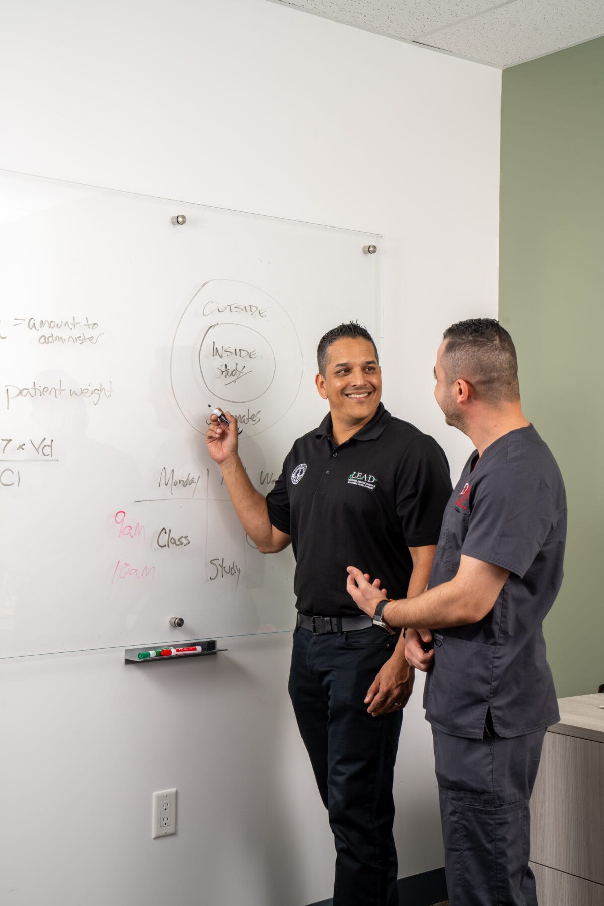 Two men stand by a whiteboard discussing a diagram with labeled circles; one points at the board while the other faces him, both appear engaged in conversation.