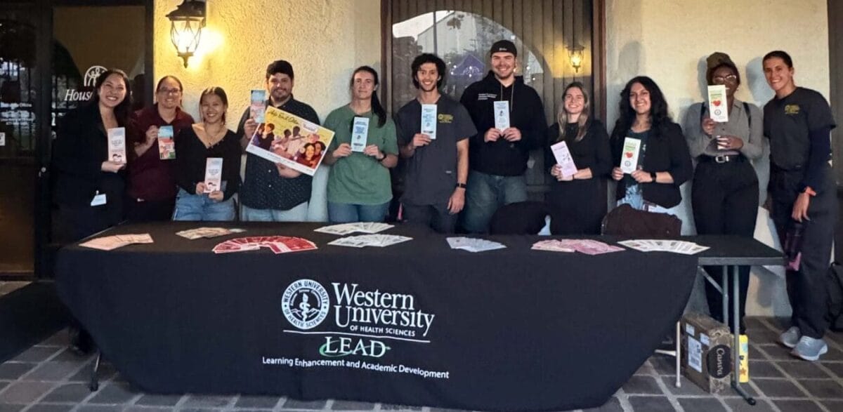 A group of people stands behind a table with a "Western University LEAD" tablecloth, holding pamphlets and smiling, under an archway outside a building.