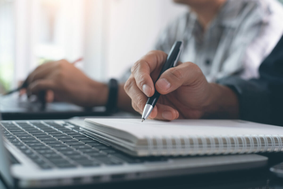 A person writing in a notebook on top of a laptop.