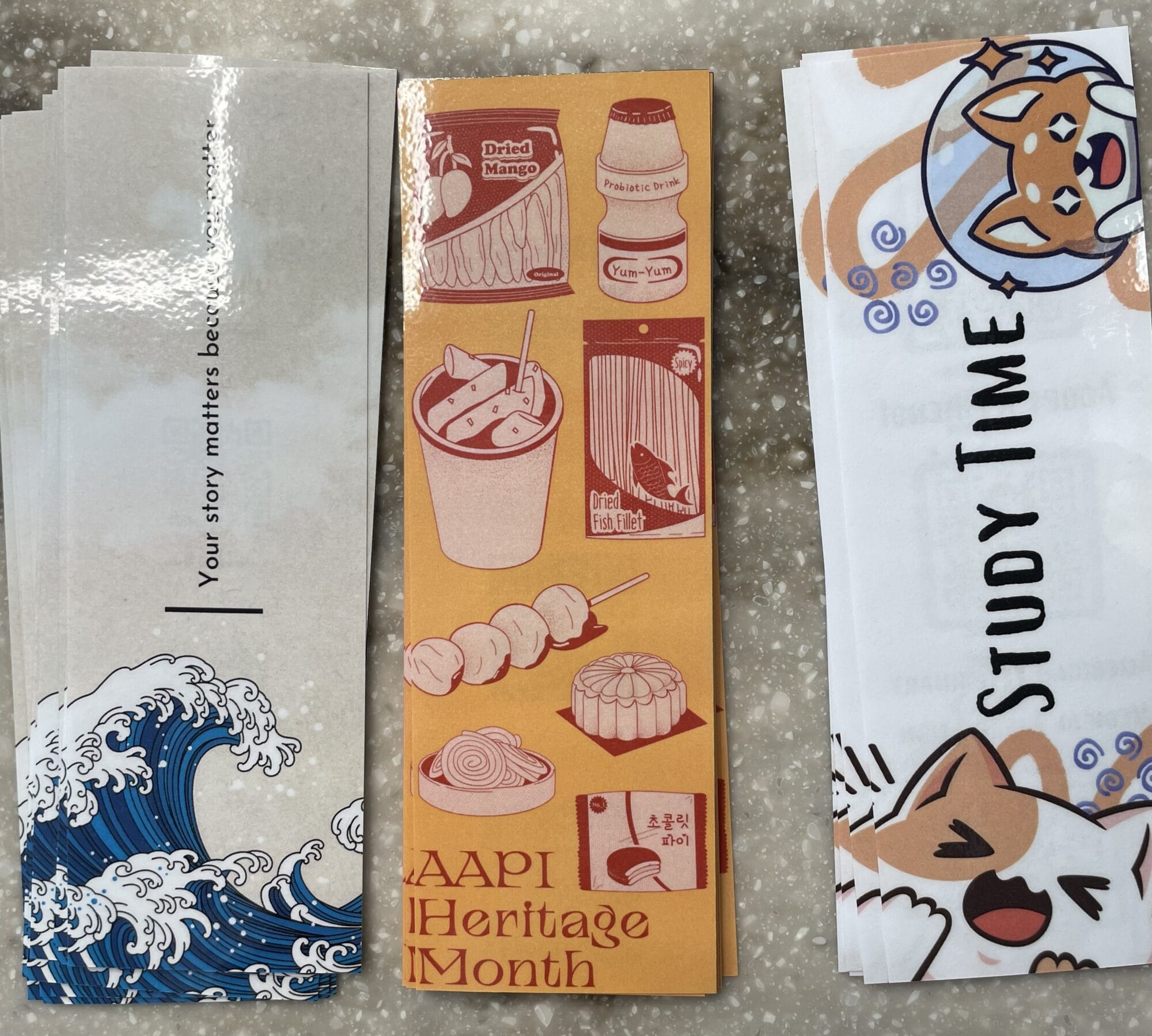 Five illustrated bookmarks arranged side by side on a marble surface, featuring waves, food, text about AAPI Heritage Month, and a cartoon fox with "STUDY TIME" text.