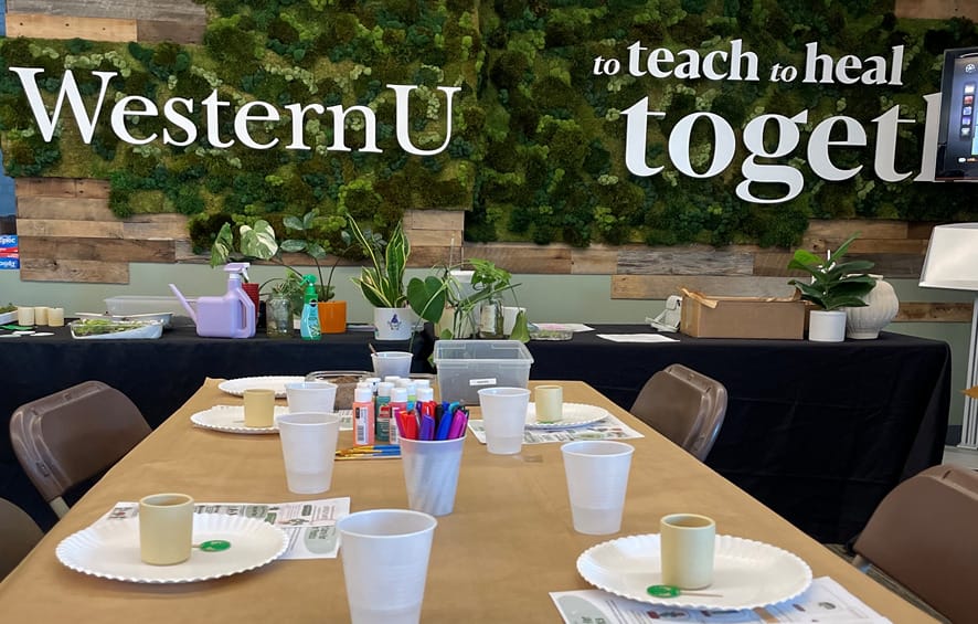 Table set with art supplies and small containers in front of a green wall displaying the words "WesternU to teach to heal toget.
