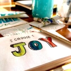 A sketchbook page with the words “CHOOSE JOY” in colorful letters, a paintbrush on top, and art supplies and paper scattered around.