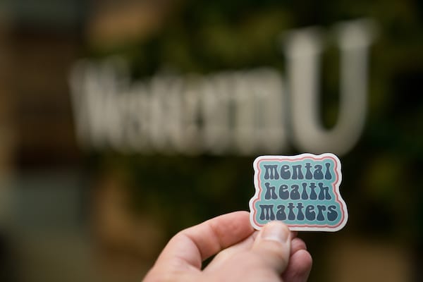 A hand holds a sticker reading "mental health matters" with a blurred "Western U" sign in the background.