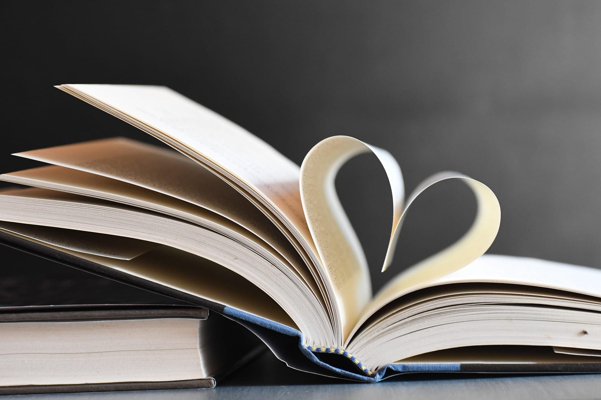An open book with two pages curved to form a heart shape, resting on top of a closed book against a plain background.
