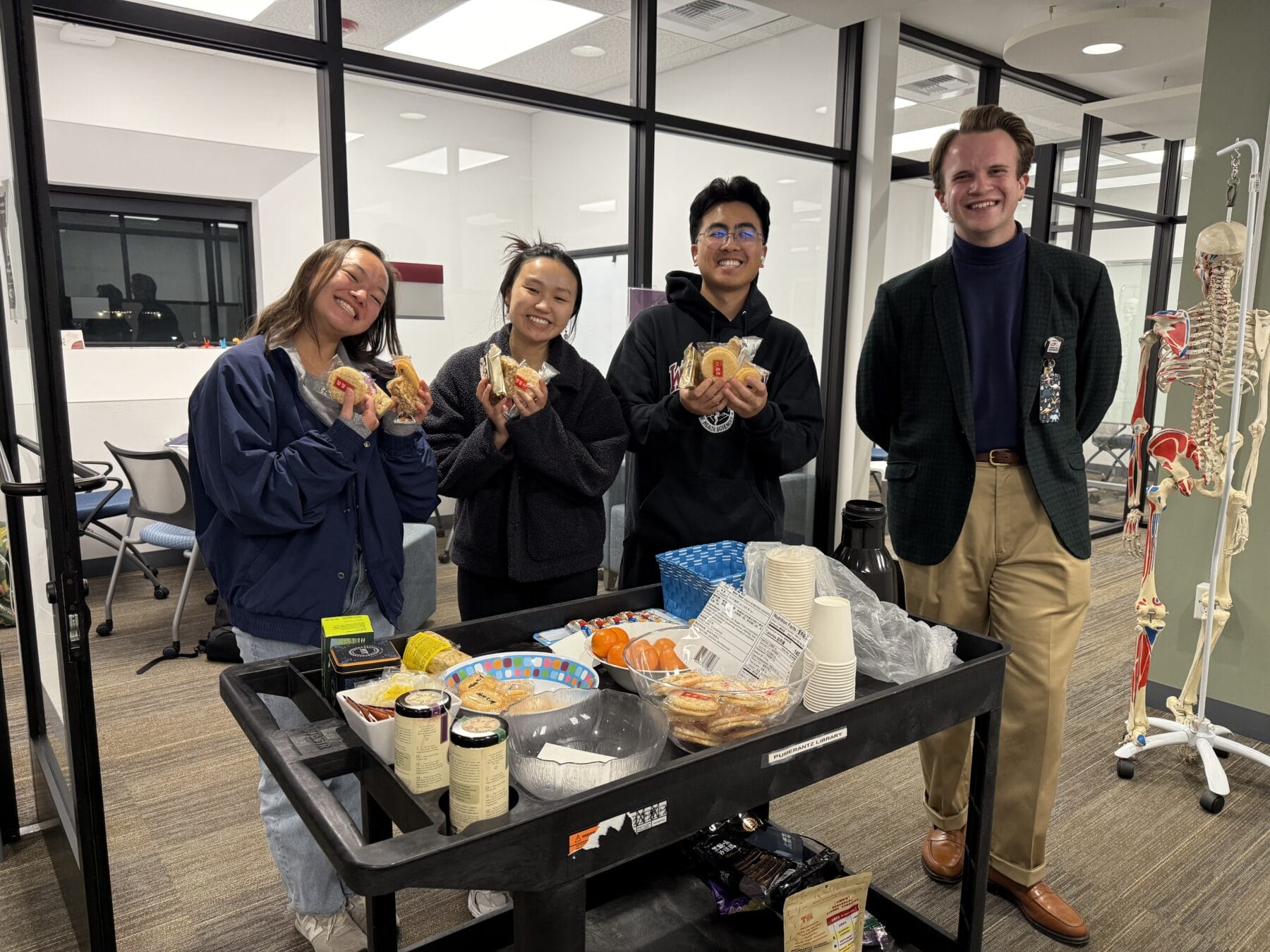 Four people stand by a cart with food and drinks in an office setting. Three hold sandwiches and smile, while one stands with hands clasped. A skeleton model is visible in the background.