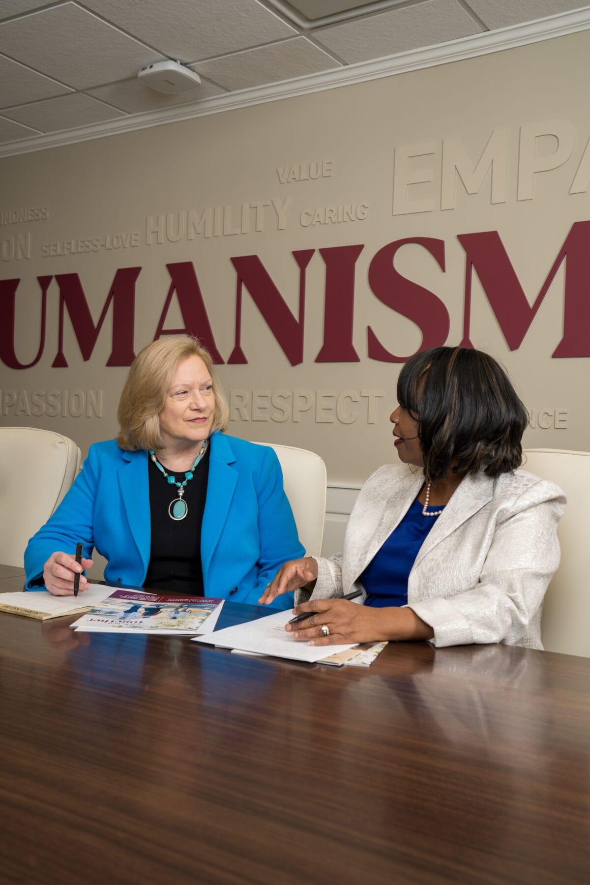 Two women sit at a conference table in discussion, with documents in front of them and the word "HUMANISM" on the wall behind them.