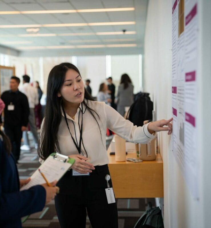 A woman in a professional setting points to a poster on the wall while another person follows along with a clipboard. Various individuals can be seen in the background of the well-lit room.