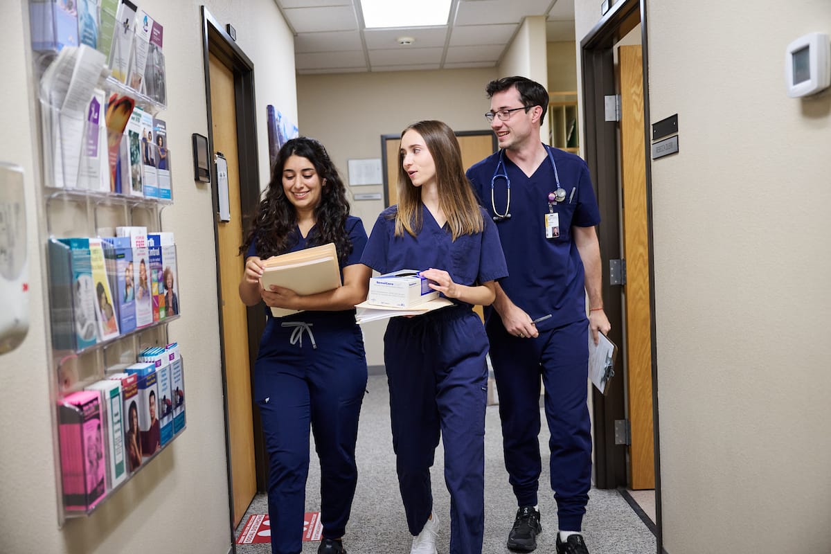 Three healthcare professionals in scrubs walk down a hallway; one holds patient files, another carries a box, and the third has a stethoscope around his neck.
