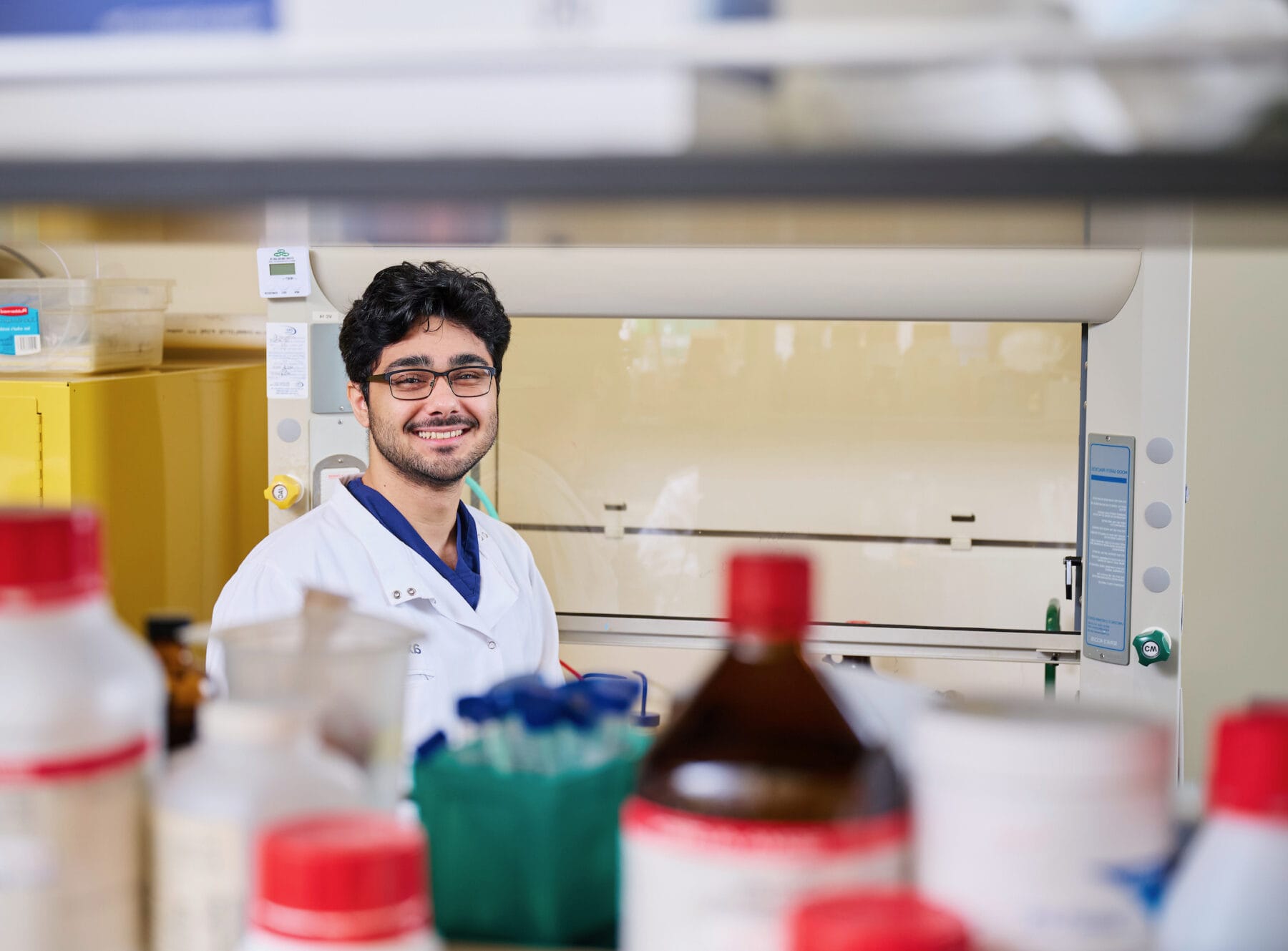 A person wearing a lab coat and glasses is smiling in a laboratory, surrounded by various bottles and lab equipment.
