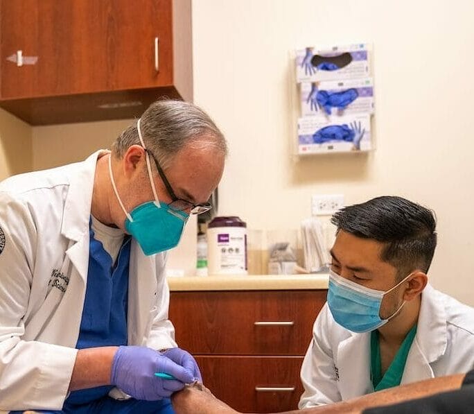 Two doctors wearing masks examine a patient's foot in a clinical setting. One doctor is holding the patient's foot while the other provides assistance. Medical supplies and cabinets are visible in the background.