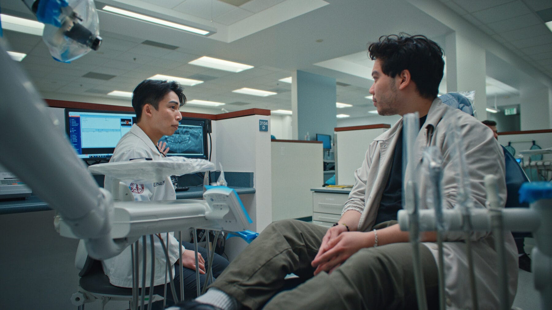 A dentist consults with a seated patient in a modern dental clinic, surrounded by dental equipment and a computer displaying an X-ray.