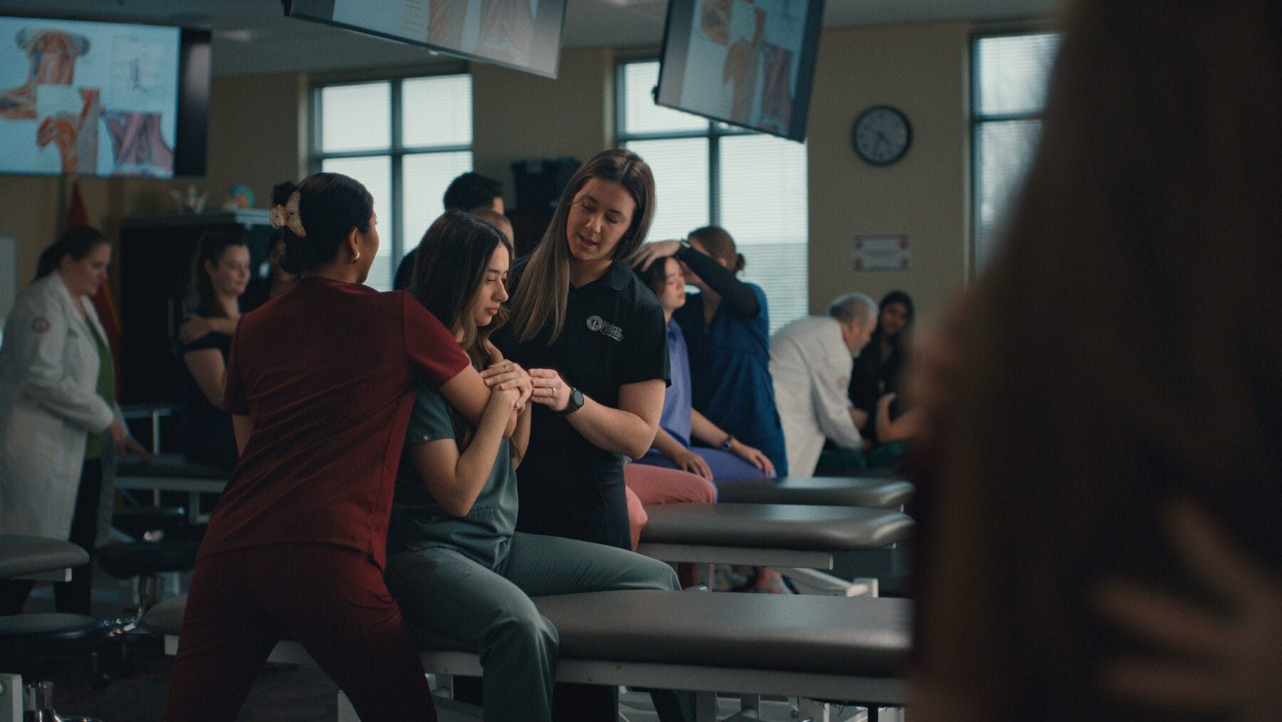 A group of students practice physical therapy techniques in a classroom, with two students assisting a seated classmate on a treatment table.