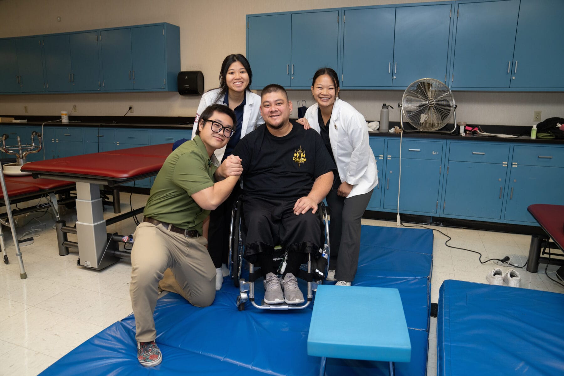 A group of four people are in a room with blue cabinets. One person is seated in a wheelchair, and the others are standing or kneeling beside him, all smiling.