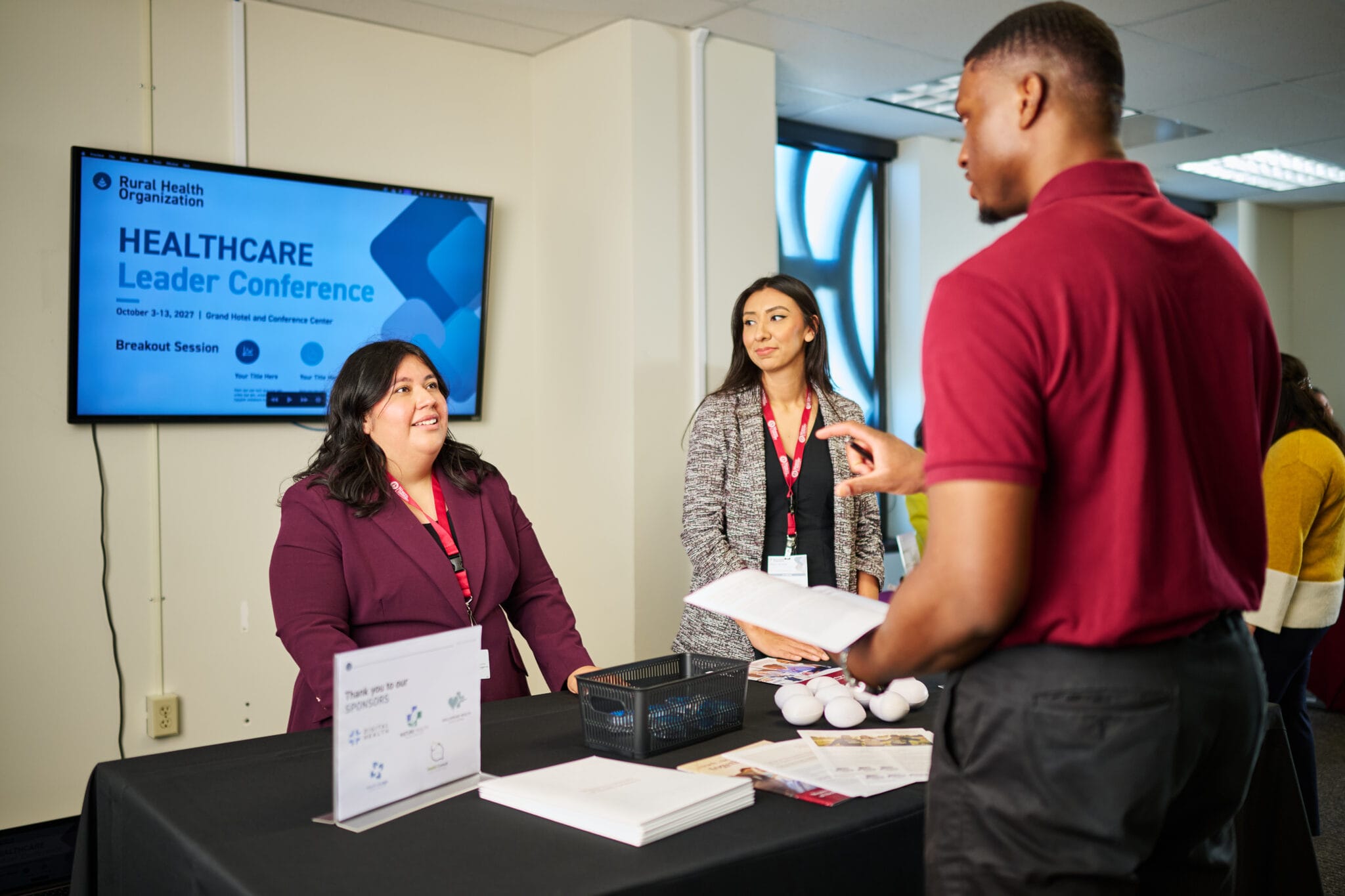 Three people converse at an information booth during a Healthcare Leader Conference, with a display screen and promotional materials visible on the table.