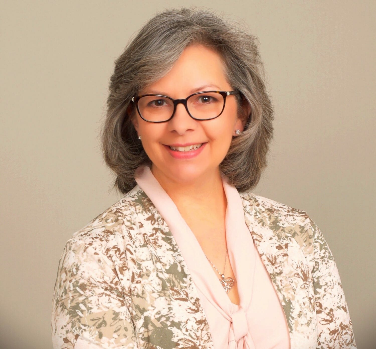 A person with glasses and gray hair smiles, wearing a patterned jacket over a light blouse against a plain background.