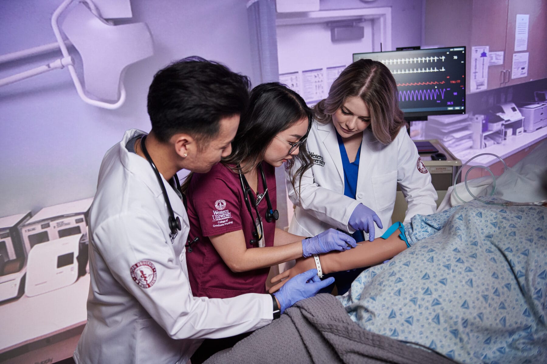 Three medical students practice drawing blood on a simulation mannequin in a clinical skills lab with medical equipment in the background.