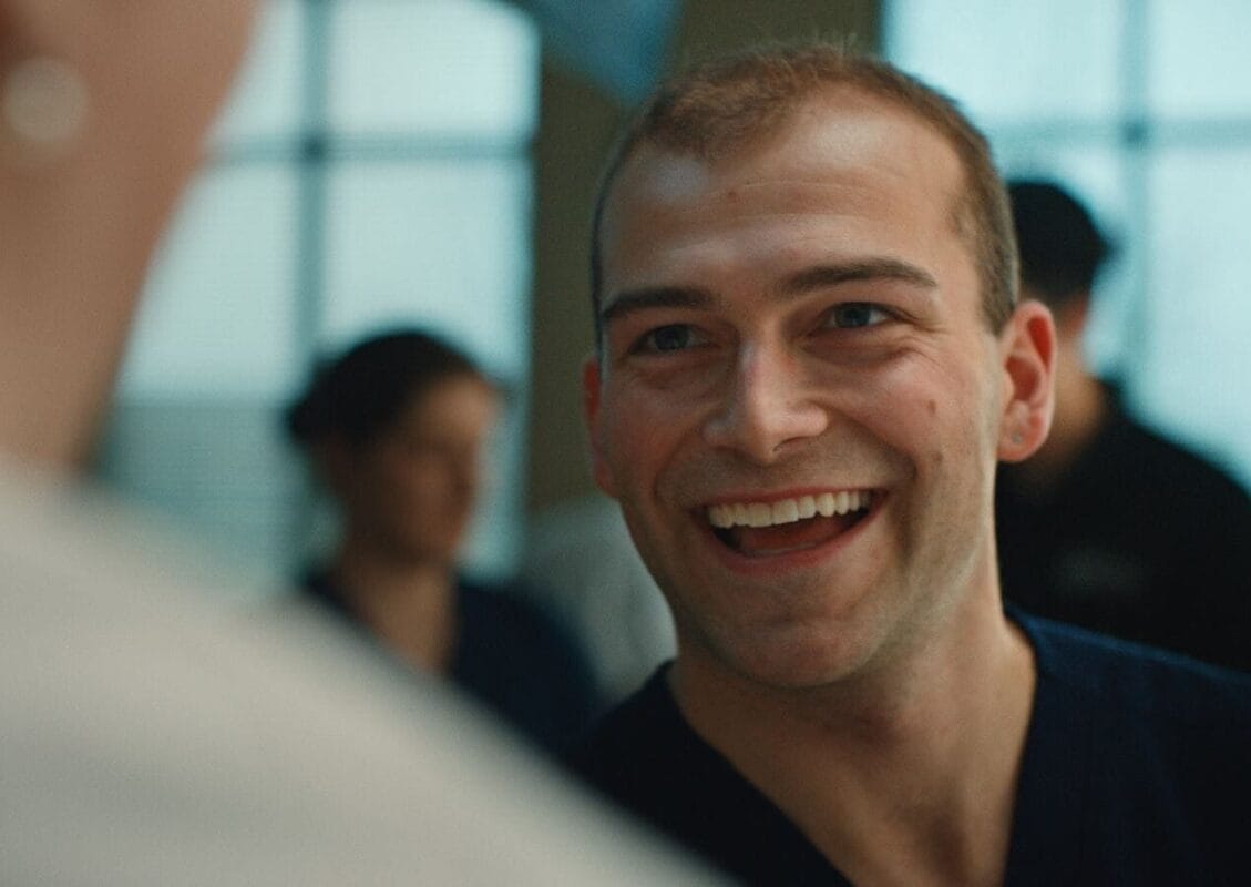 A man in scrubs smiles and looks at someone off-camera in a brightly lit room with blurred people in the background.