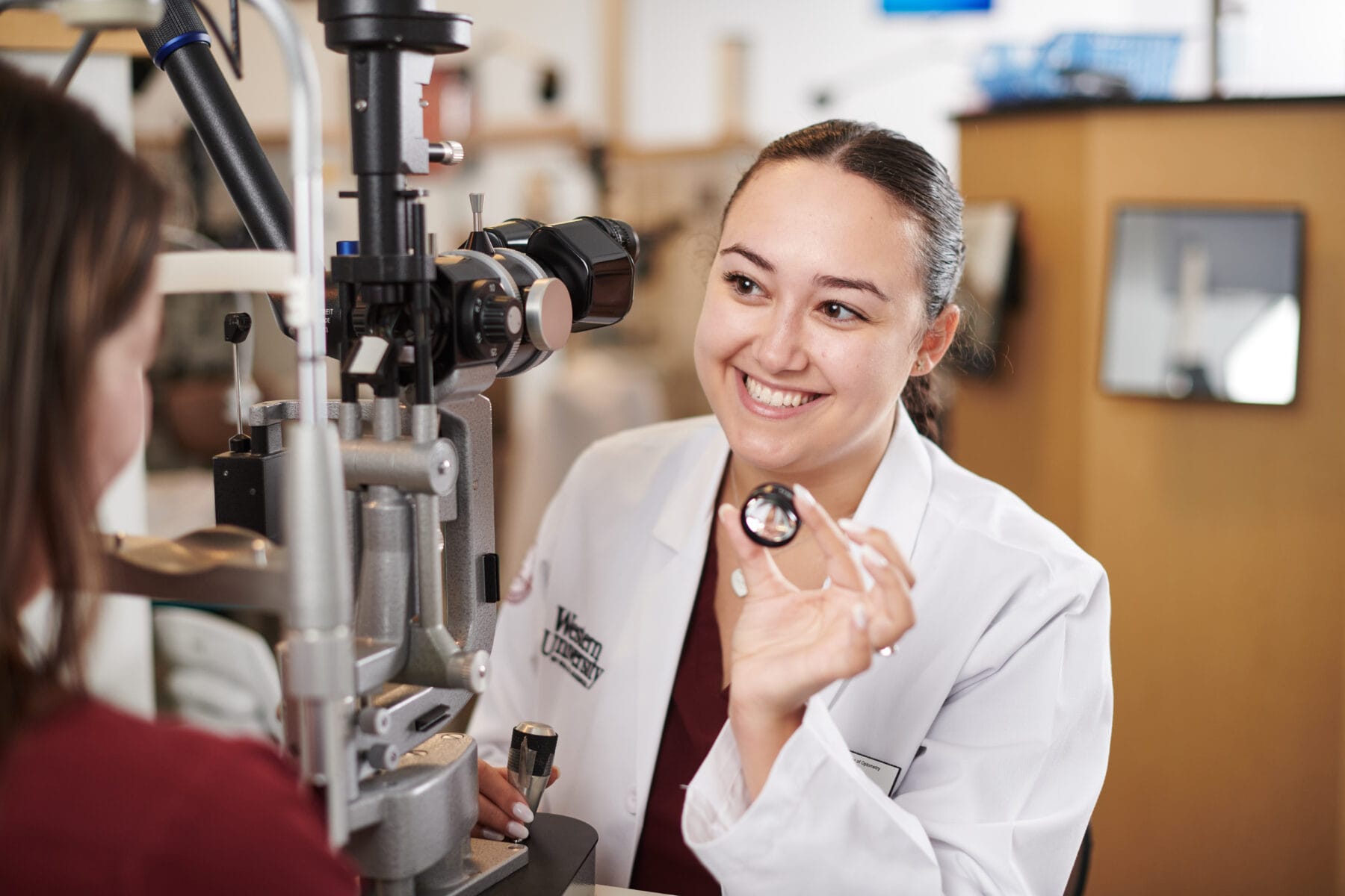 A female optometrist in a white coat holds a lens and smiles while preparing to examine a patient’s eyes using ophthalmic equipment.