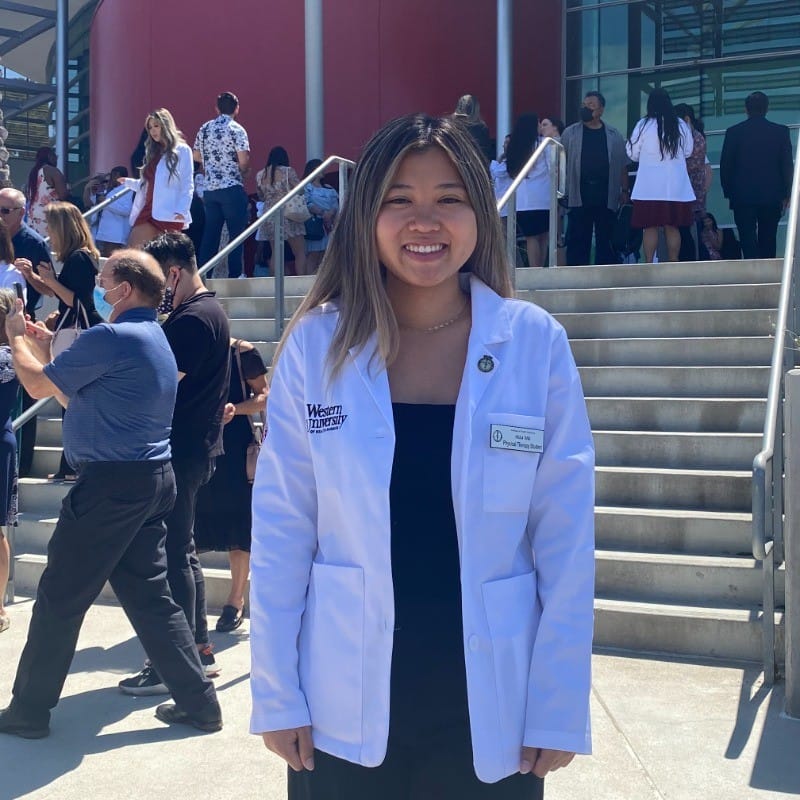 A person in a white coat stands in front of steps, smiling. People are gathered and taking photos in the background.