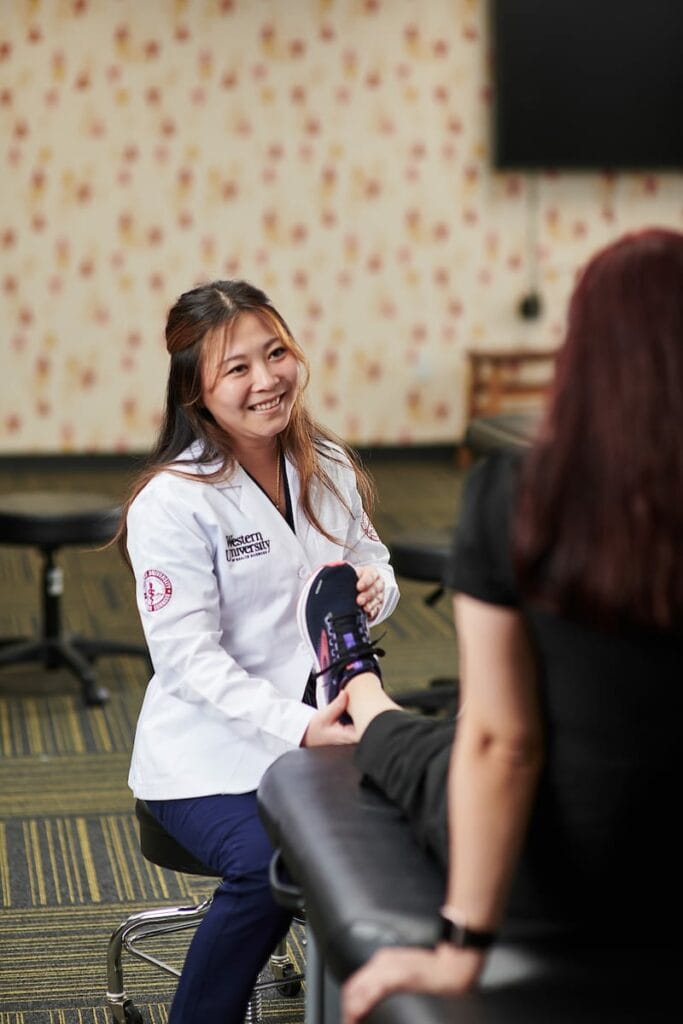 A female healthcare professional in a white coat performs a foot examination on a seated patient inside a medical office.