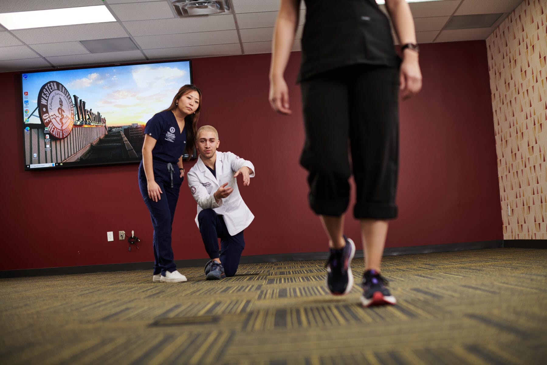 Two healthcare professionals observe and assess a person walking in a clinical room with a red wall and patterned carpet.