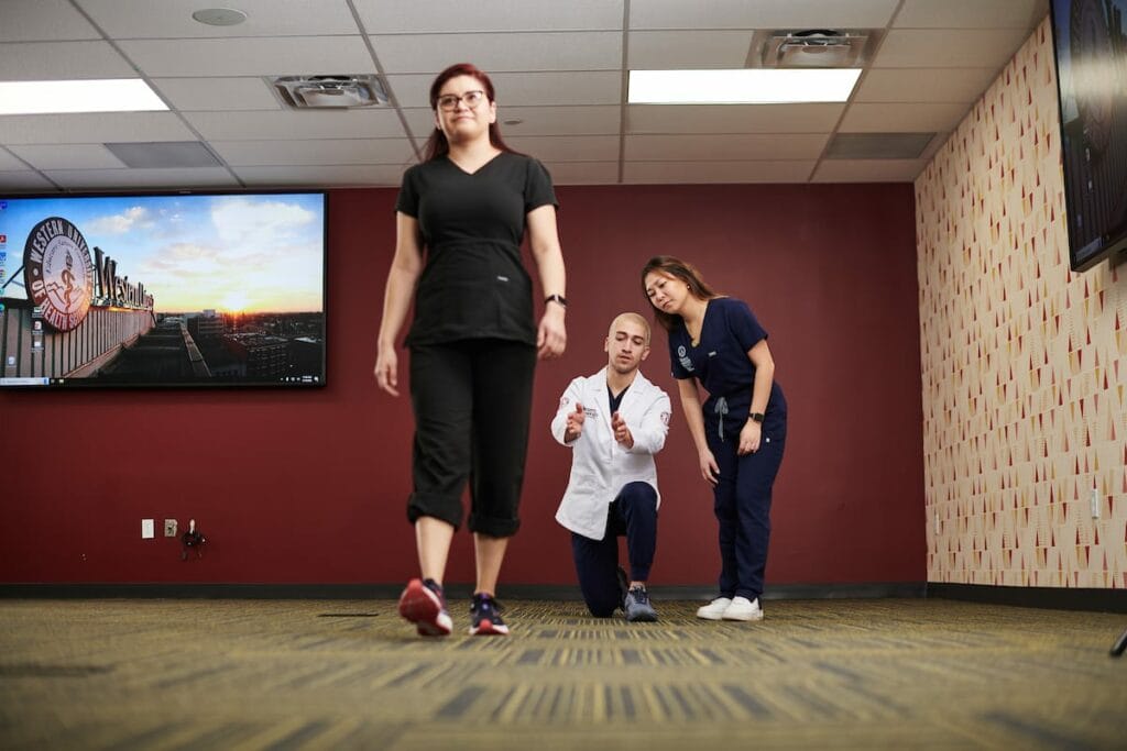 A person in black walks while a man in a white coat kneels and points, and a woman in blue scrubs watches in a room with a wall-mounted screen.