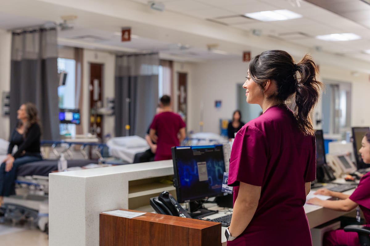 A nurse in maroon scrubs stands at a hospital workstation, while another nurse works at a computer and a patient talks with staff in the background.