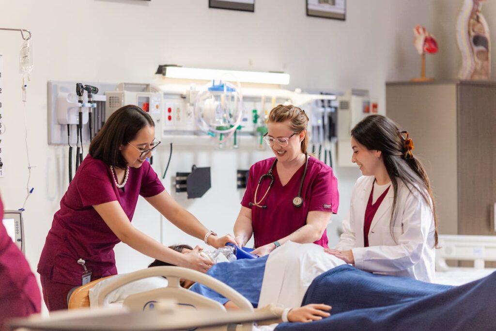Three healthcare professionals work together beside a hospital bed, attending to a patient mannequin in a medical simulation lab.
