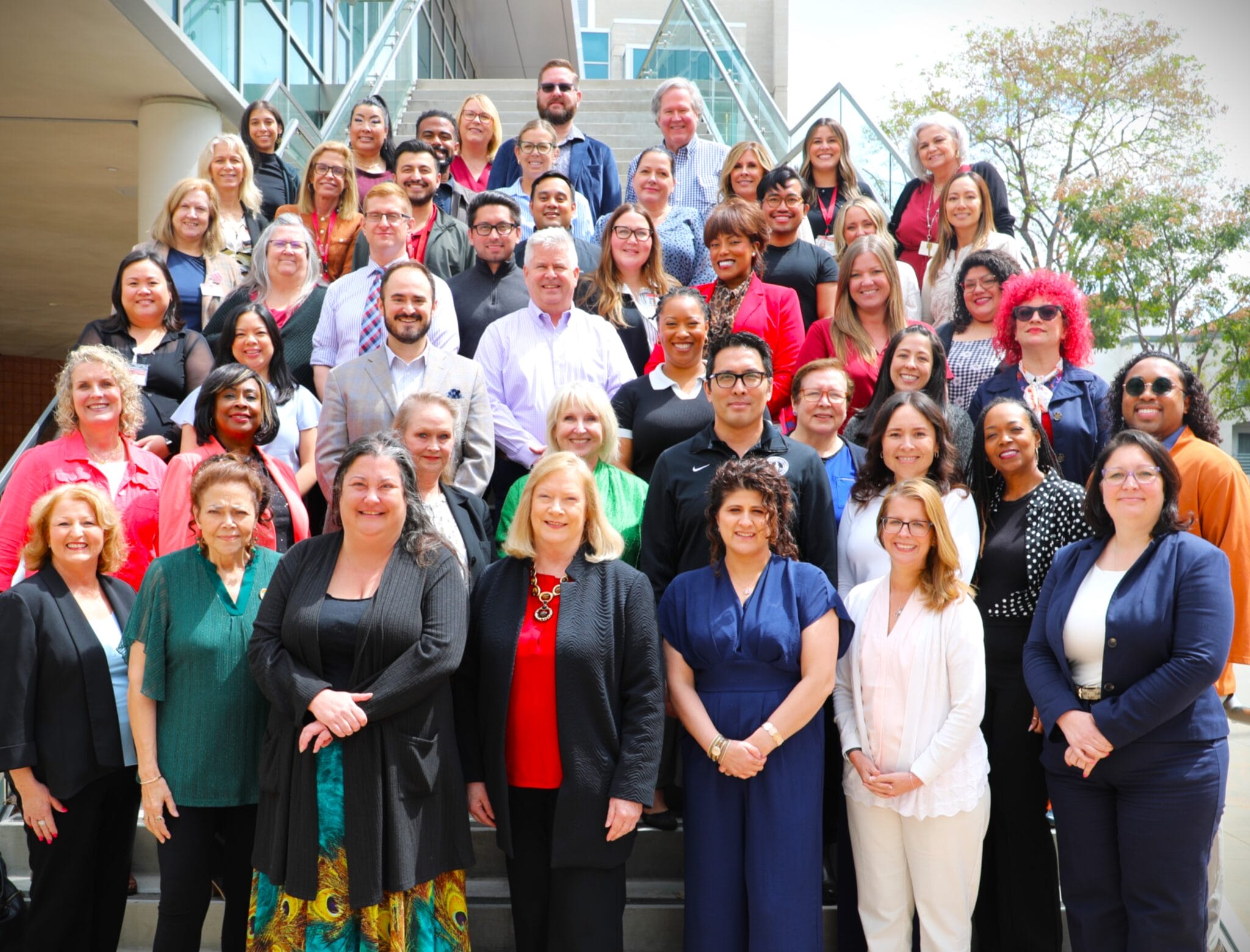 A large group of professionally dressed people pose together on stair steps outside a building, smiling at the camera.