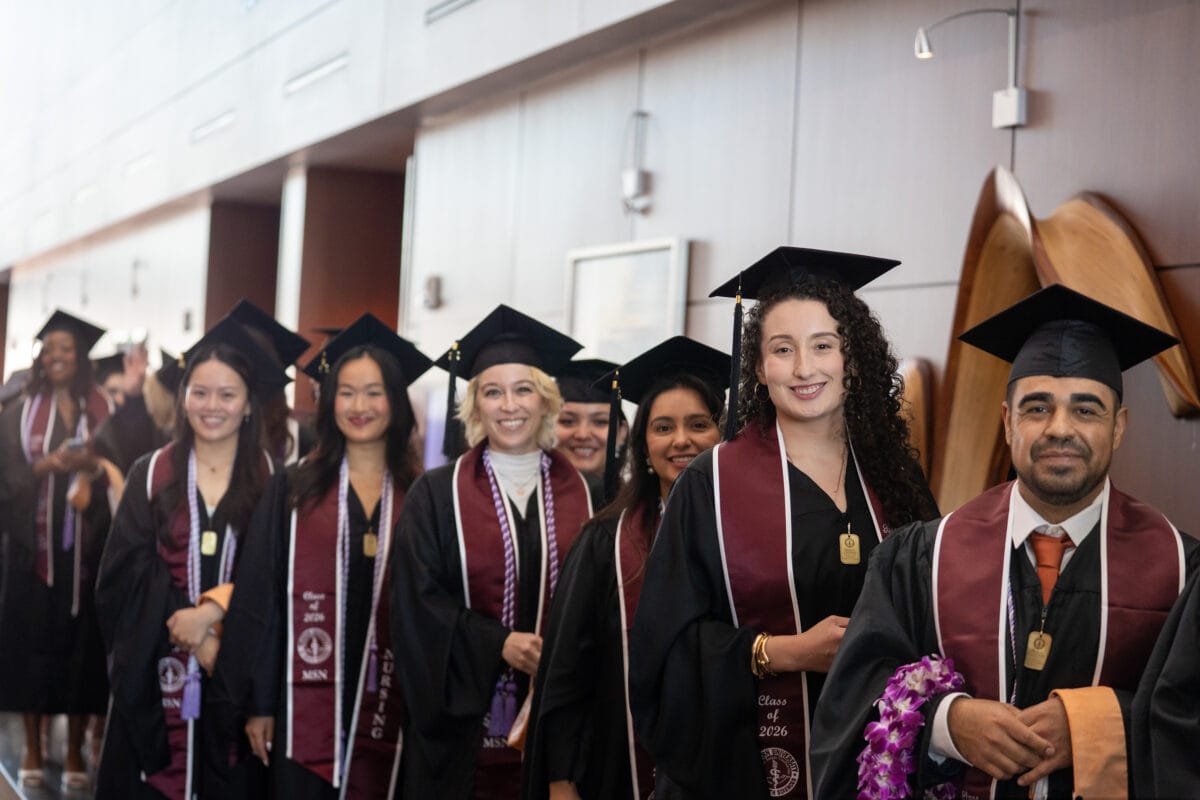 Graduates in caps and gowns stand in a line indoors, smiling and waiting for a ceremony to begin.