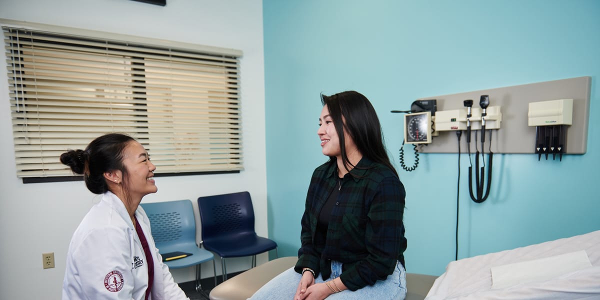 A healthcare professional talks with a patient sitting on an exam table in a medical office, with diagnostic tools on the wall behind them.