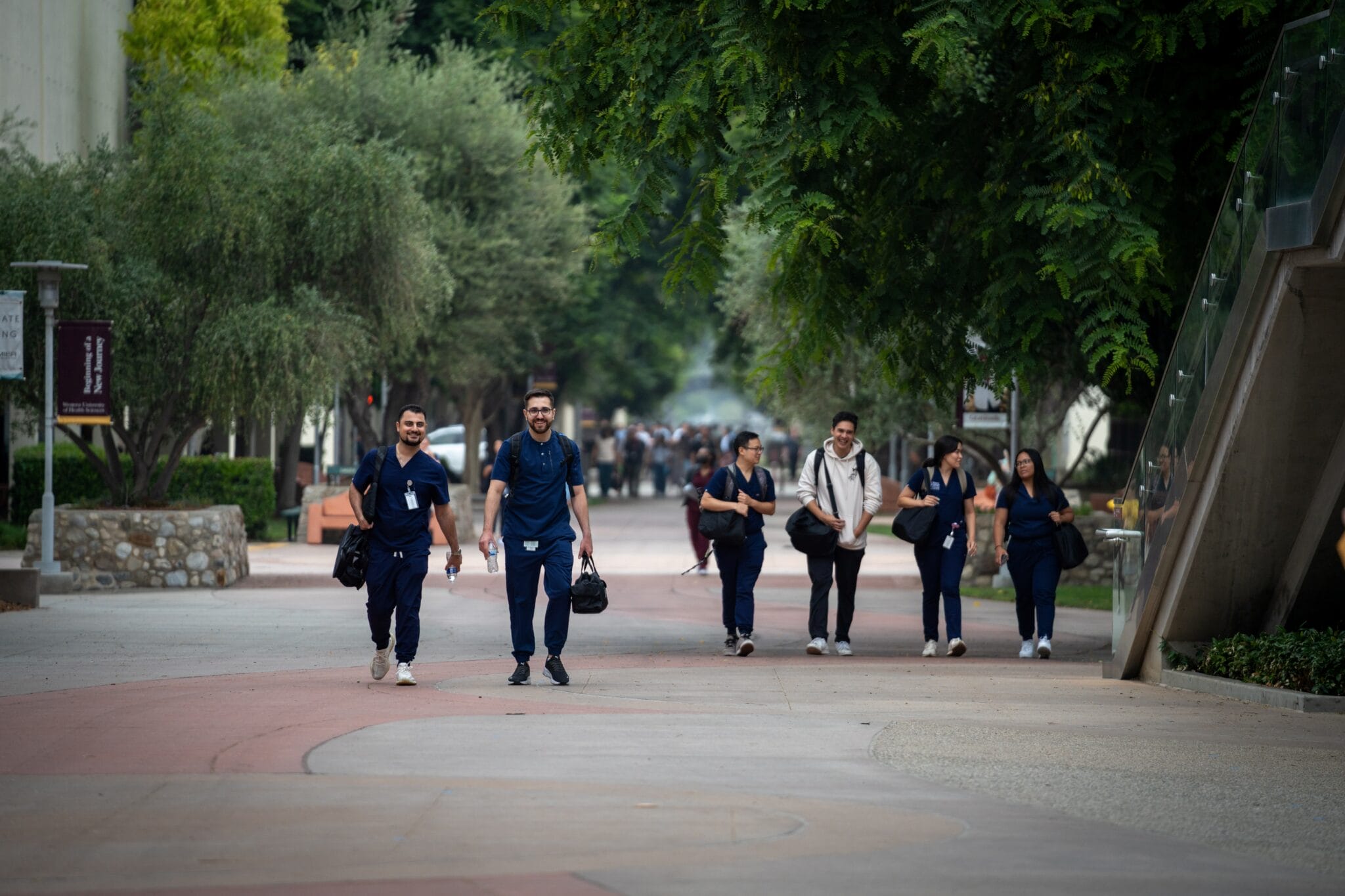 A group of people in scrubs walk along a tree-lined campus pathway, carrying bags and talking with each other.