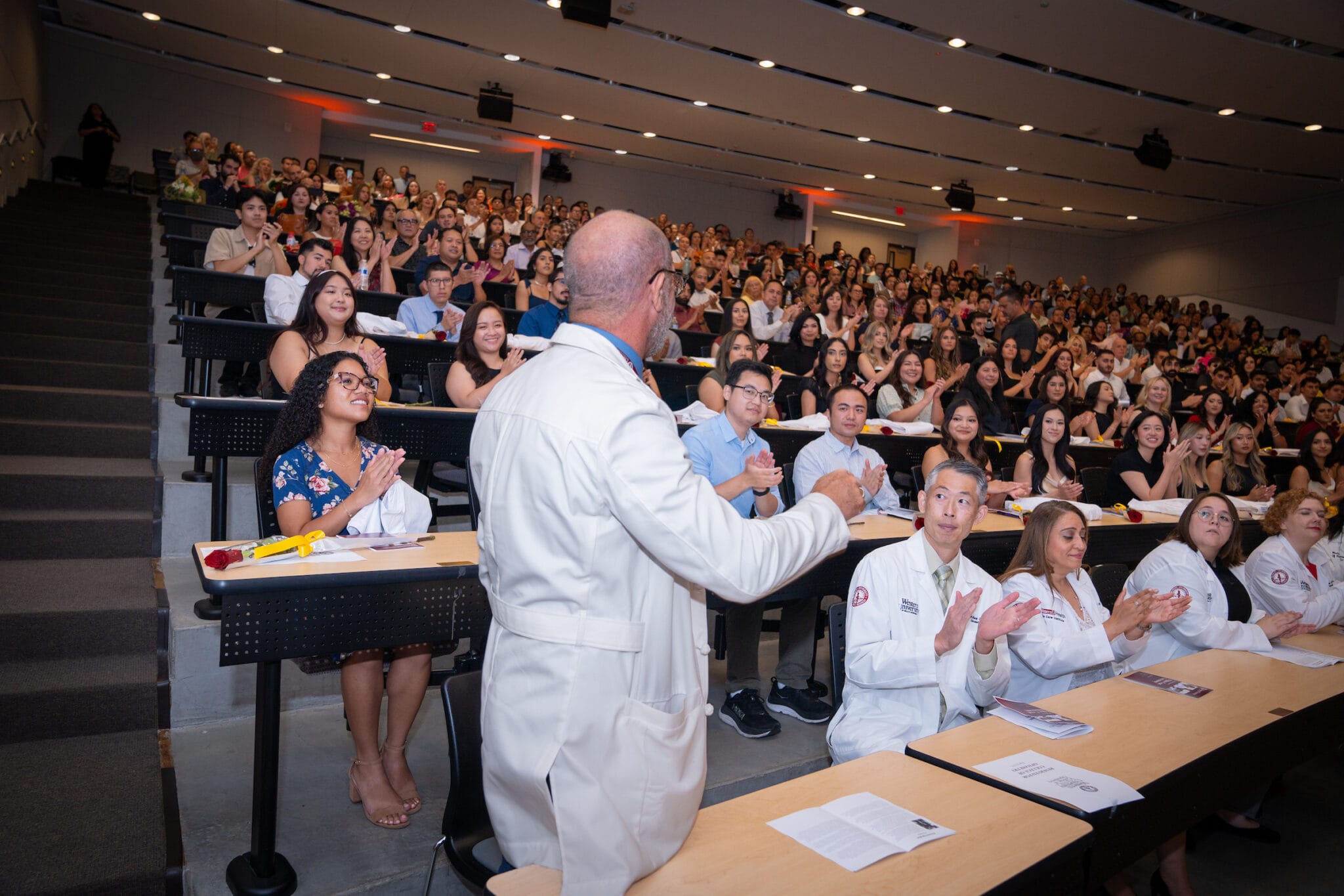 A person in a white coat addresses a packed lecture hall, with attendees sitting and clapping, some also wearing white coats.