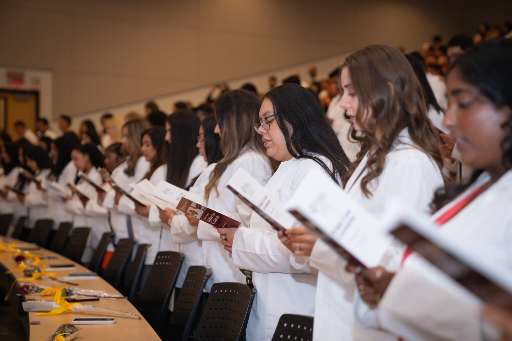 A group of people in white coats stand in rows, holding papers and reading during a ceremony in a large lecture hall.