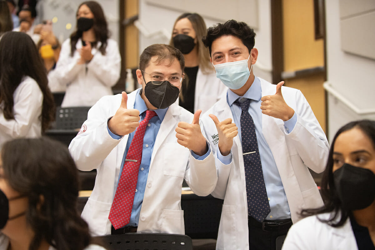 Two students in white coats, in classroom giving thumbs up