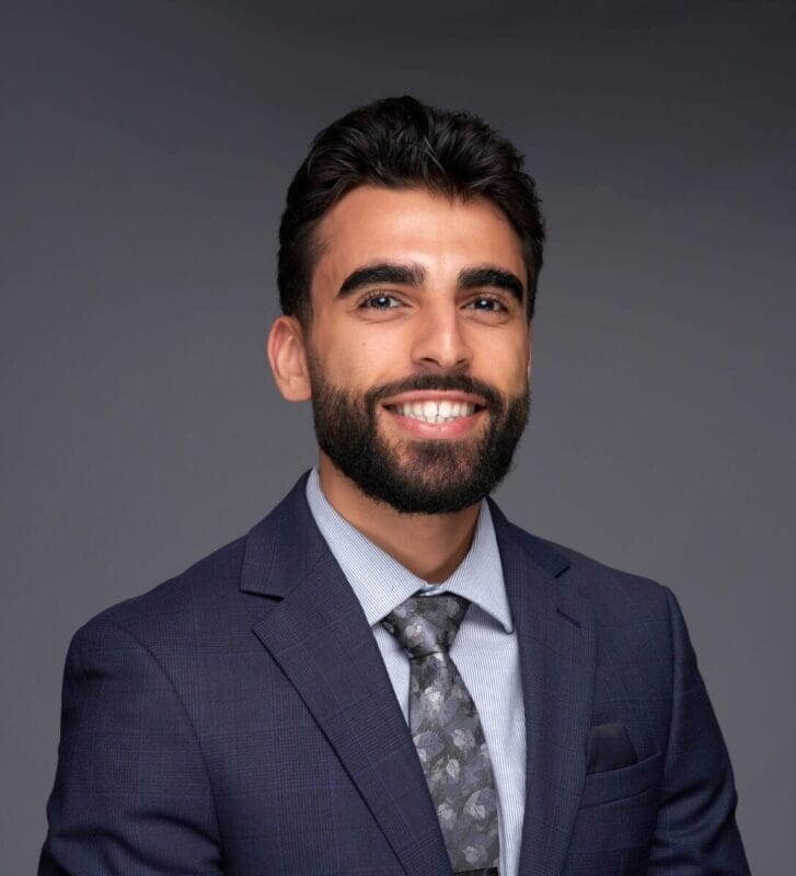 Man with dark hair and beard wearing a blue suit, light shirt, and patterned tie, smiling against a plain gray background.