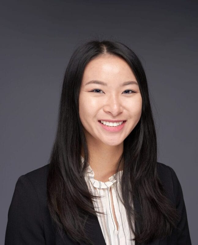 A woman with long, straight black hair, wearing a black blazer and light blouse, smiles at the camera against a plain grey background.