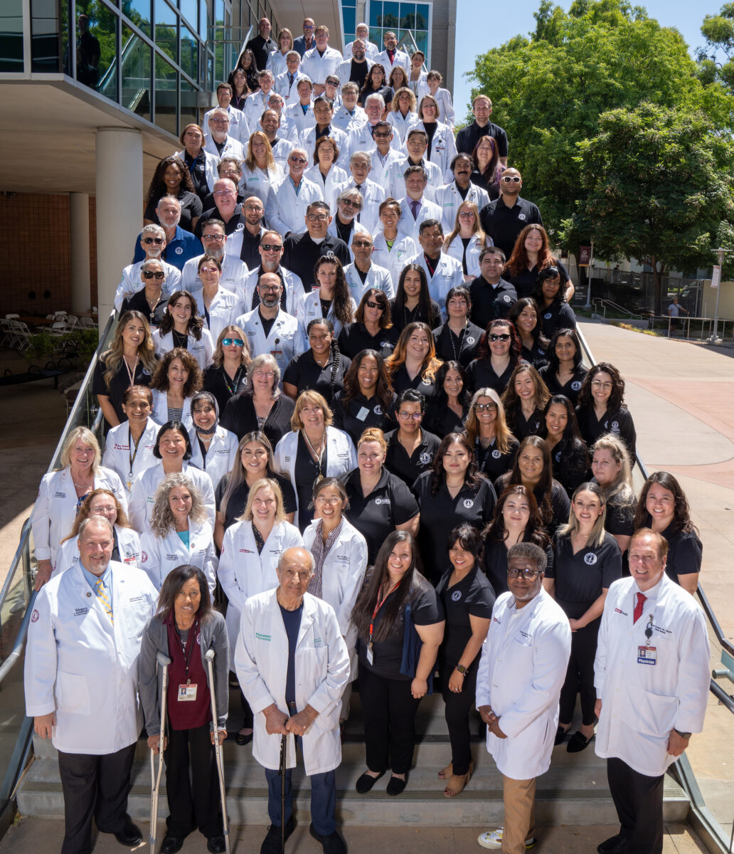 Group photo of diverse medical staff with doctors, nurses and administrators on outdoor steps, dressed in lab coats and professional attire.