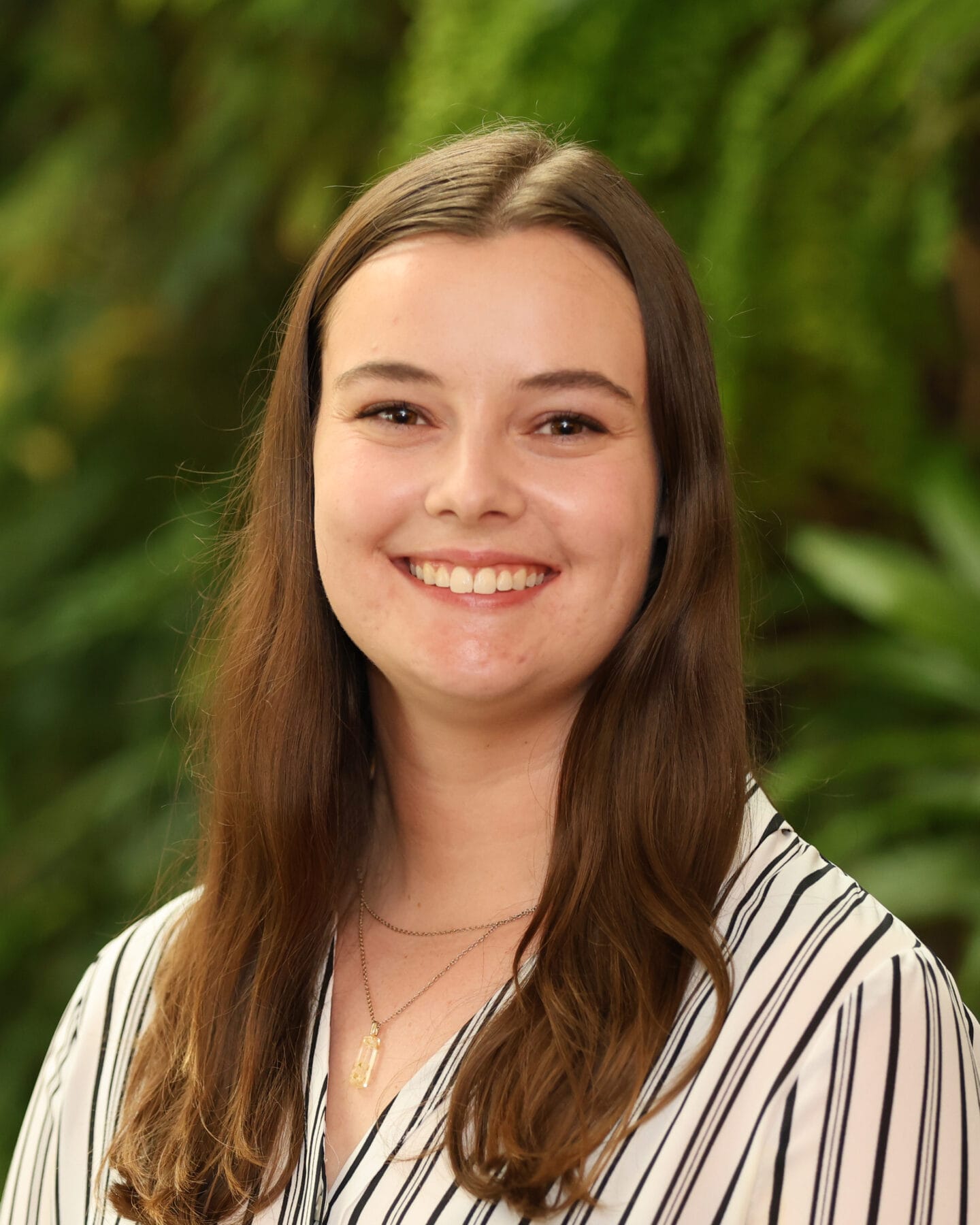 A young woman with long brown hair, wearing a striped blouse and gold necklace, smiles at the camera with greenery in the background.