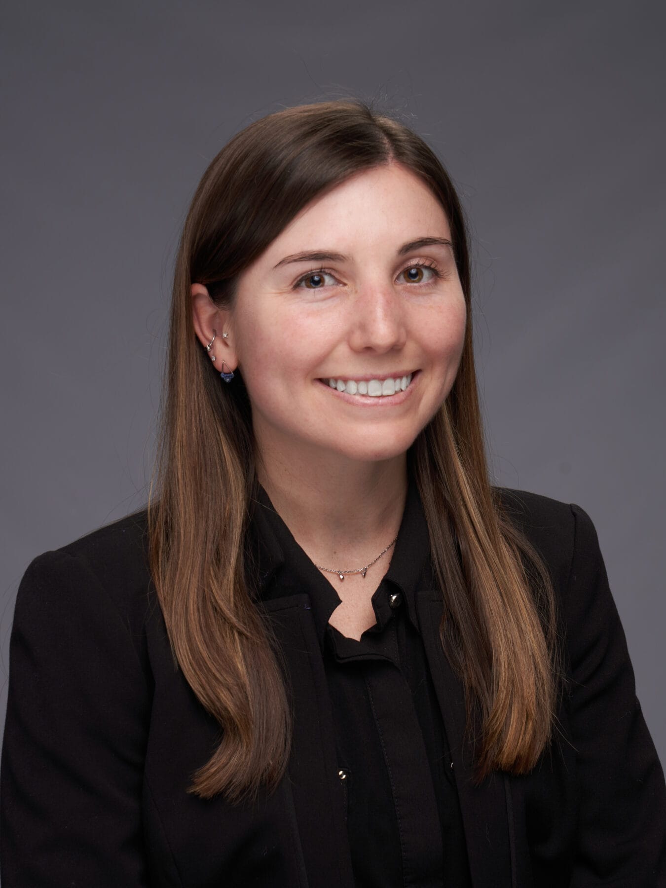 A woman with straight brown hair, wearing a black blazer and black shirt, smiles at the camera against a plain gray background.