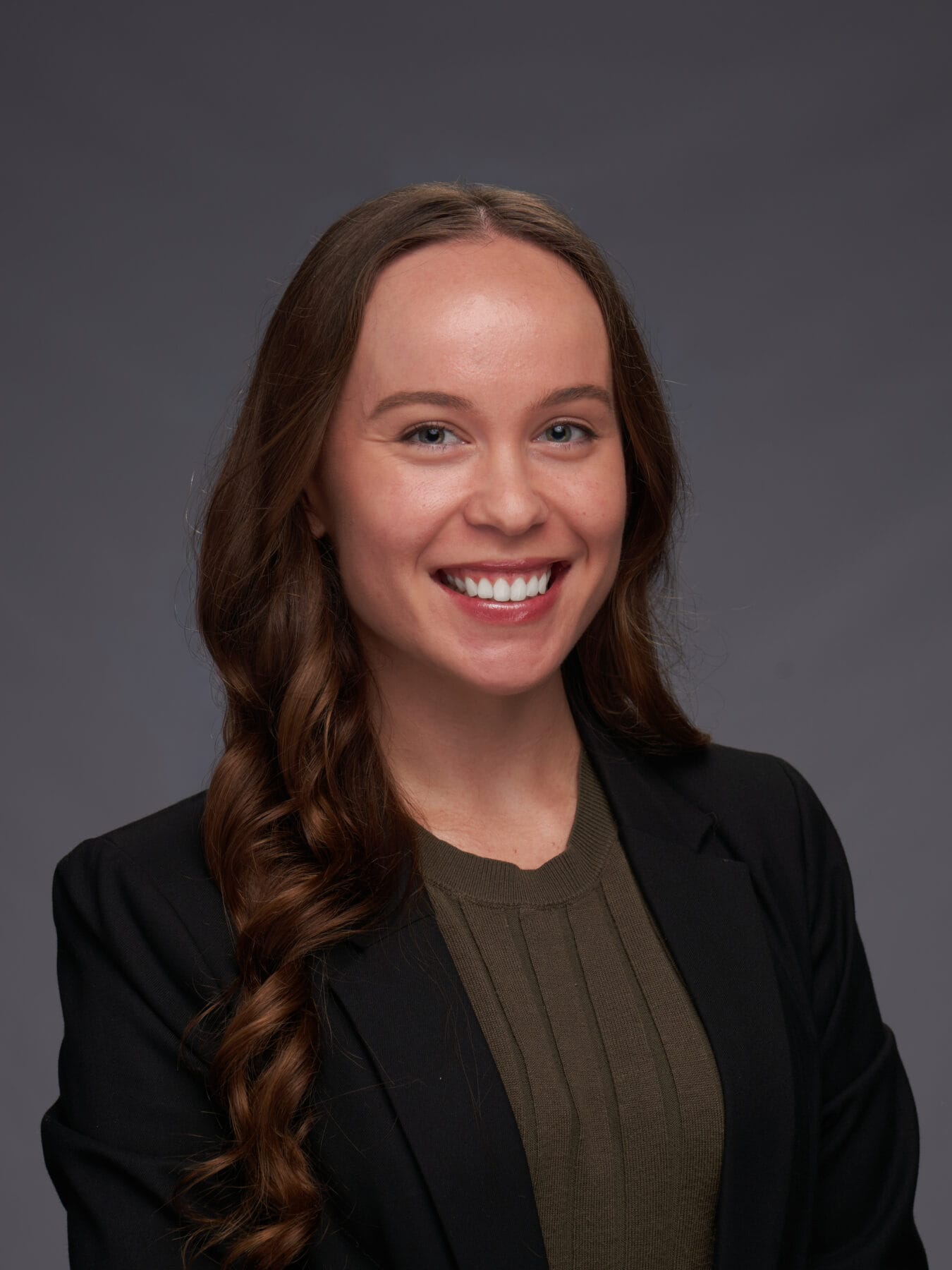 A woman with long brown hair, wearing a dark blazer and olive green top, smiles in front of a plain gray background.