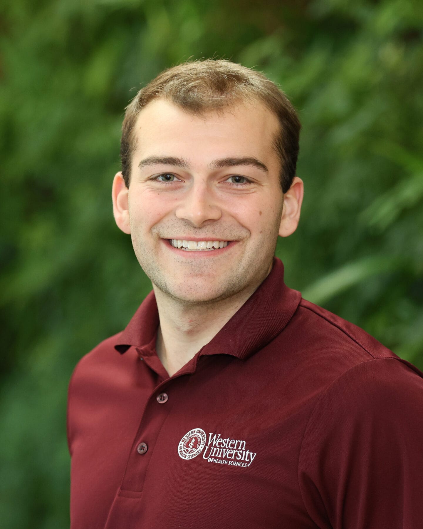 A smiling person wearing a maroon Western University Health Sciences polo shirt poses in front of a green, blurred background.