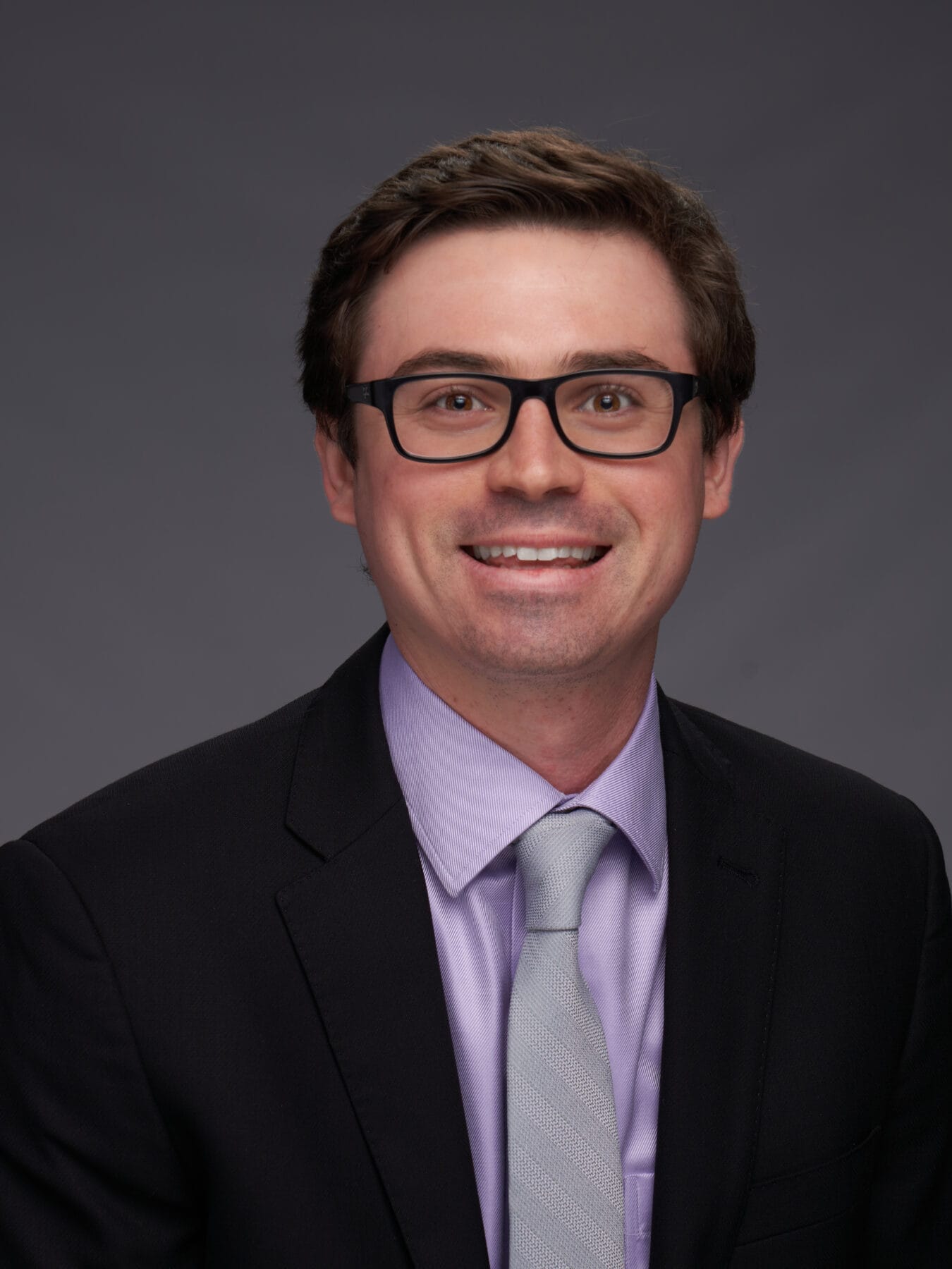 A man with short brown hair and glasses, wearing a dark suit, light purple shirt, and light gray tie, smiles at the camera against a plain gray background.