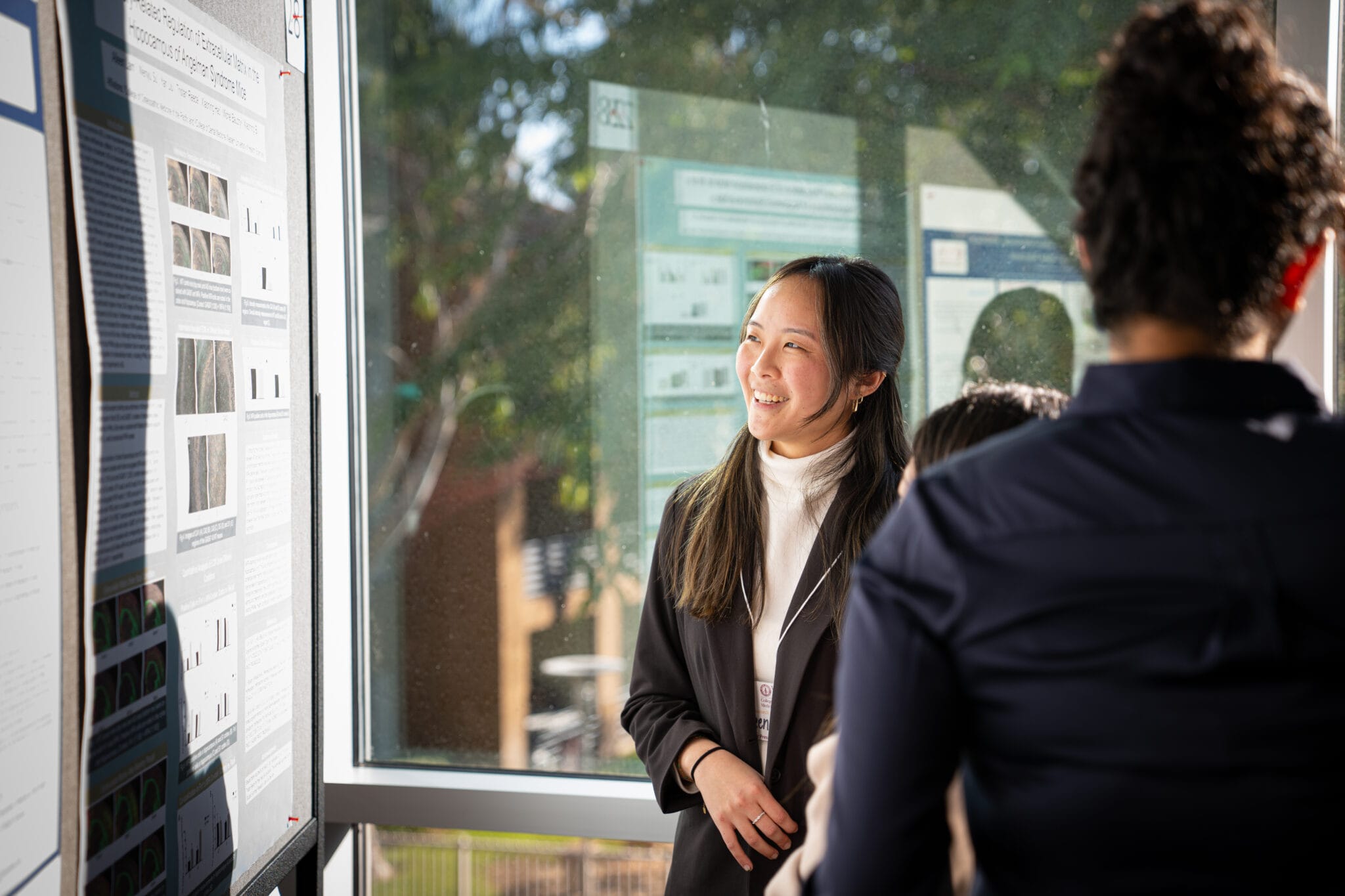 A woman in business attire stands by a poster presentation, smiling and talking to two people indoors near large windows.