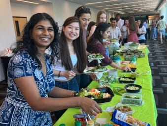 People gather around a long table covered with green tablecloths, serving themselves food from various trays and containers at a casual indoor event.