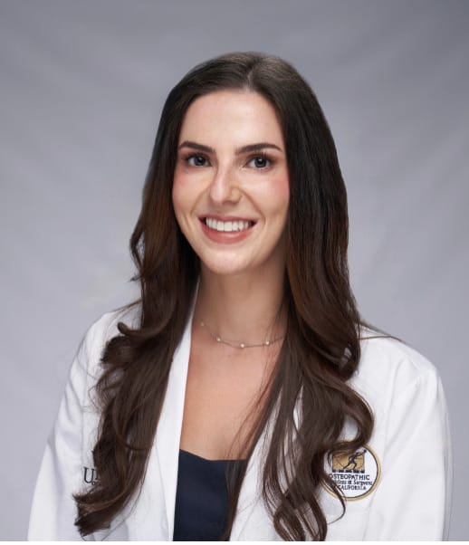 Woman with long brown hair wearing a white medical coat and navy top, smiling against a plain light gray background.