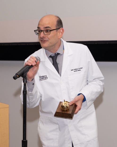 A man in a white lab coat speaks into a microphone while holding a golden apple award plaque.