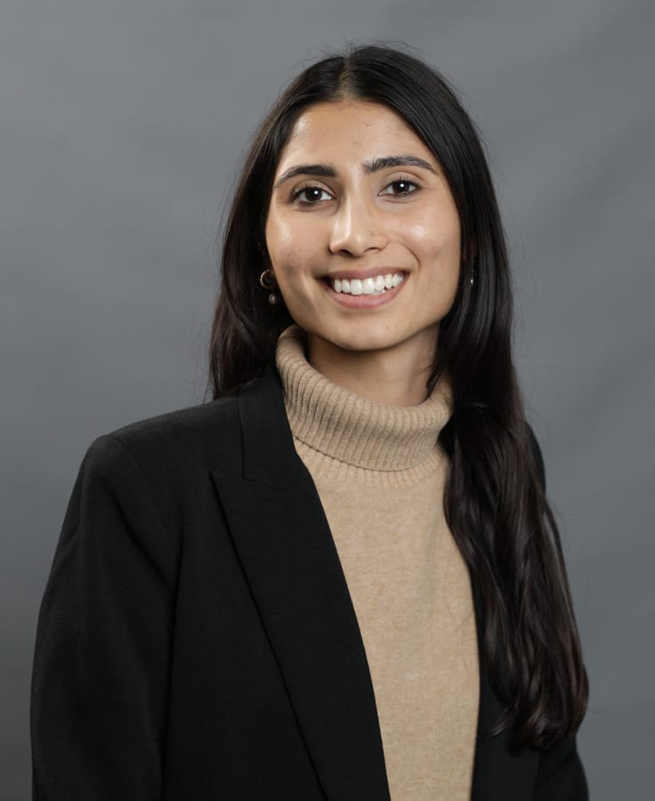 A woman with long dark hair, wearing a beige turtleneck sweater and a black blazer, poses for a studio portrait against a plain grey background.