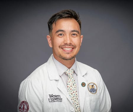 A man wearing a white lab coat and tie poses for a formal portrait against a gray background. The lab coat displays "Western University" and several pins and patches.