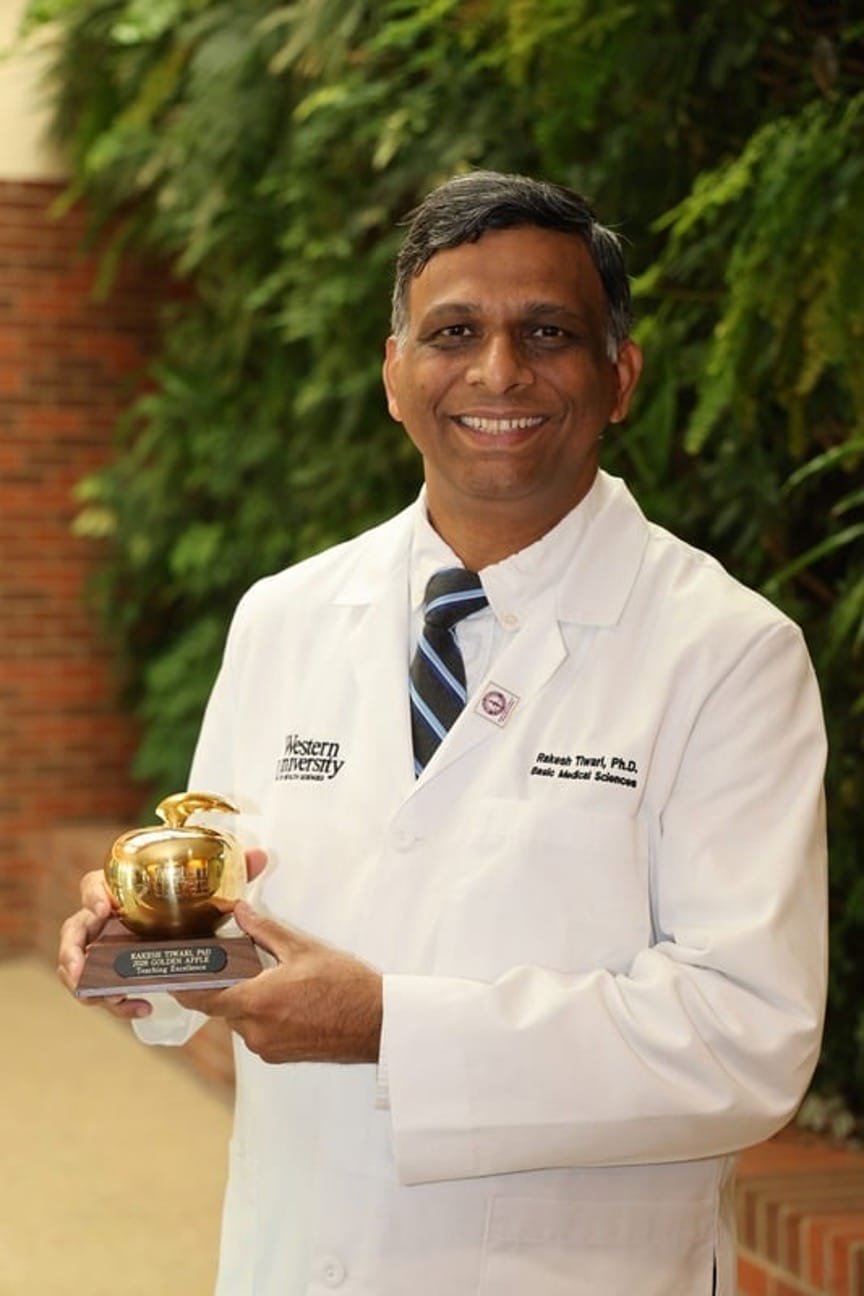 A man in a white lab coat holds a golden apple trophy and smiles at the camera. Green plants and a brick wall are in the background.